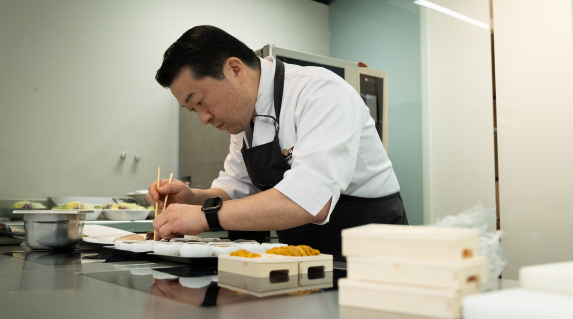 Chef professionista in divisa bianca e grembiule nero mentre prepara con cura dei piatti di sushi utilizzando le bacchette