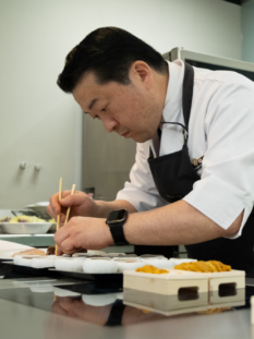 Chef professionista in divisa bianca e grembiule nero mentre prepara con cura dei piatti di sushi utilizzando le bacchette