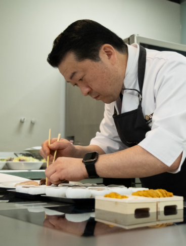 Chef professionista in divisa bianca e grembiule nero mentre prepara con cura dei piatti di sushi utilizzando le bacchette