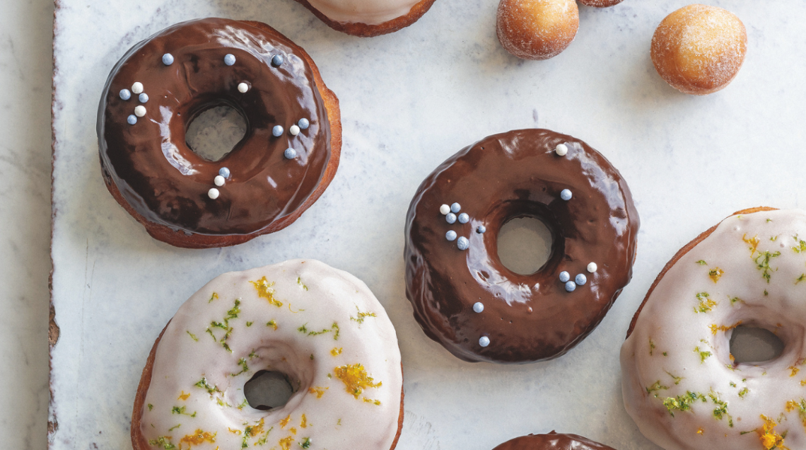 Vista dall'alto di donuts decorati con glassa al cioccolato e perline argentate, e donuts con glassa bianca e scorza di lime, su un piano di marmo