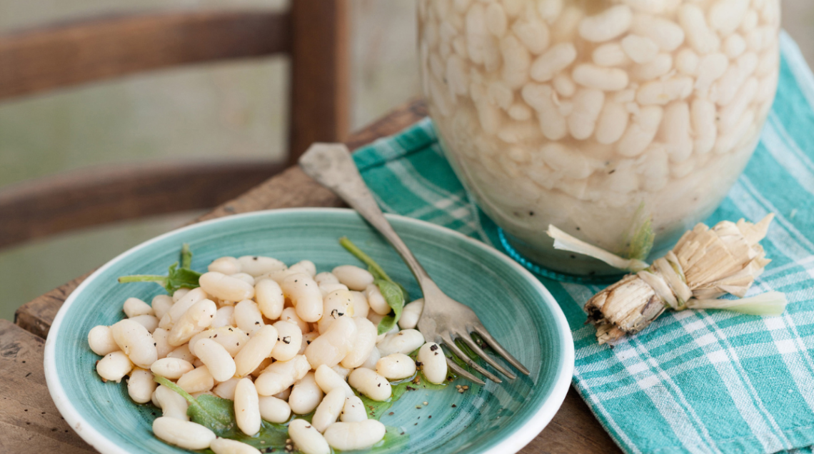Un piatto turchese con fagioli bianchi conditi con olio e pepe, accanto a un fiasco di vetro tradizionale colmo di fagioli in cottura su un tavolo rustico