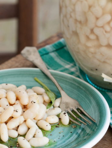 Un piatto turchese con fagioli bianchi conditi con olio e pepe, accanto a un fiasco di vetro tradizionale colmo di fagioli in cottura su un tavolo rustico
