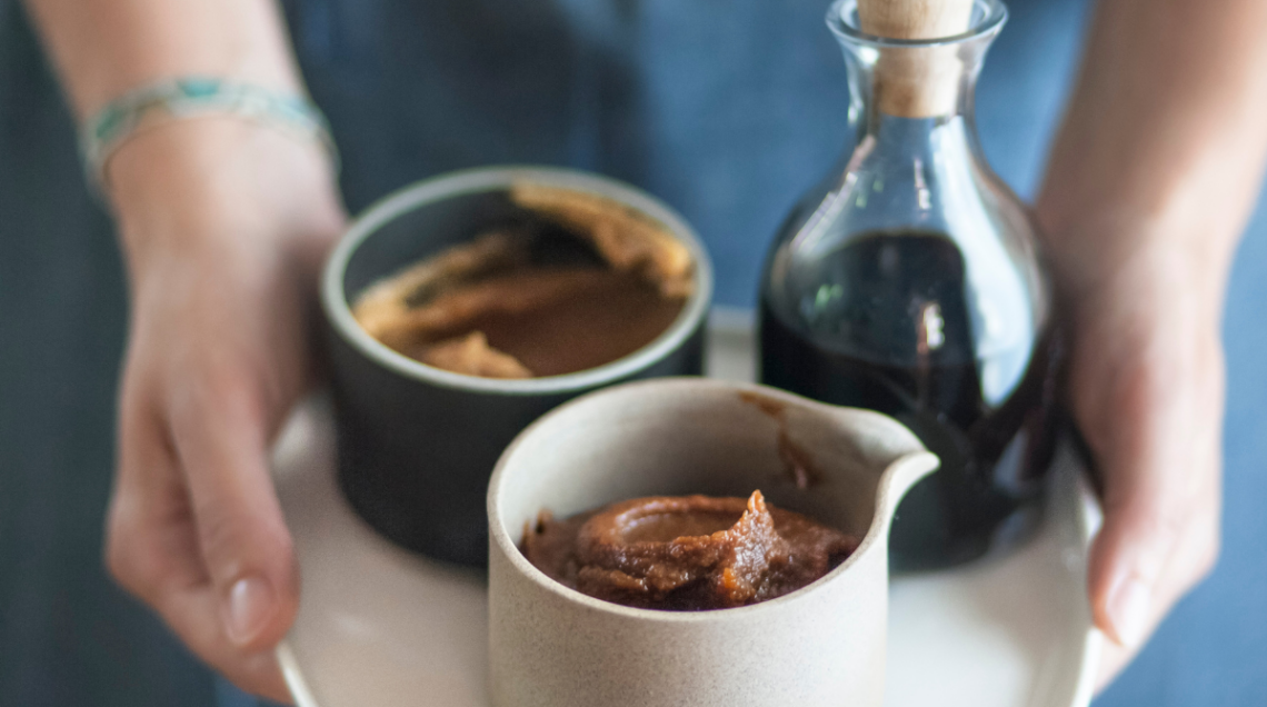 Mani che sorreggono un vassoio con due ciotole di ceramica contenenti pasta di miso e una bottiglia di vetro con salsa di soia scura