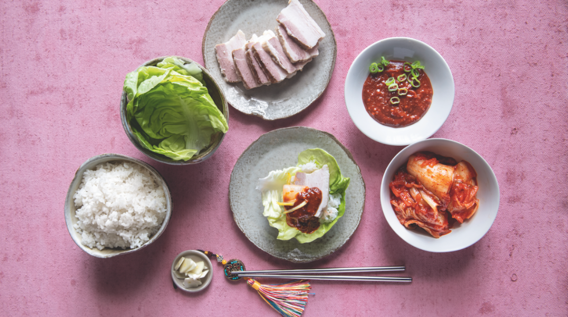 Vista dall'alto di un set da tavola coreano con ciotole di riso, kimchi, pancetta di maiale a fette, foglie di lattuga, salse piccanti e bacchette su uno sfondo rosa