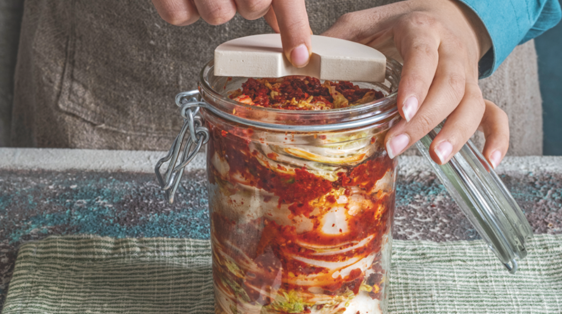Mani che pressano il cavolo cinese condito con peperoncino e spezie all'interno di un barattolo di vetro per la fermentazione, usando un peso in ceramica.