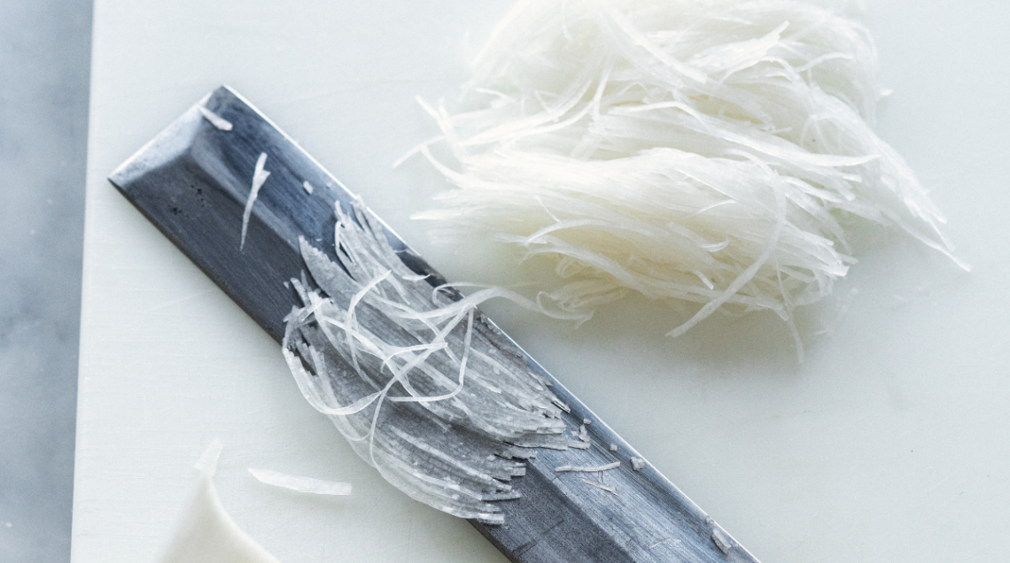 Sottili striscioline di ravanello daikon bianco appena tagliate su un tagliere, con una lama di coltello professionale in primo piano.