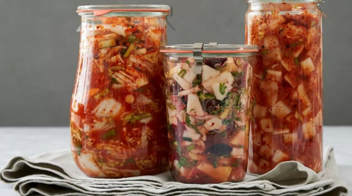 Tre barattoli di vetro di diverse dimensioni contenenti diverse varietà di kimchi fermentato, tra cui cavolo e ravanello a cubetti, su un canovaccio grigio.
