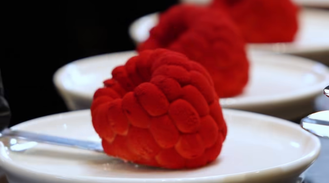 Dessert iperrealistico a forma di lampone gigante con texture granulosa rossa.