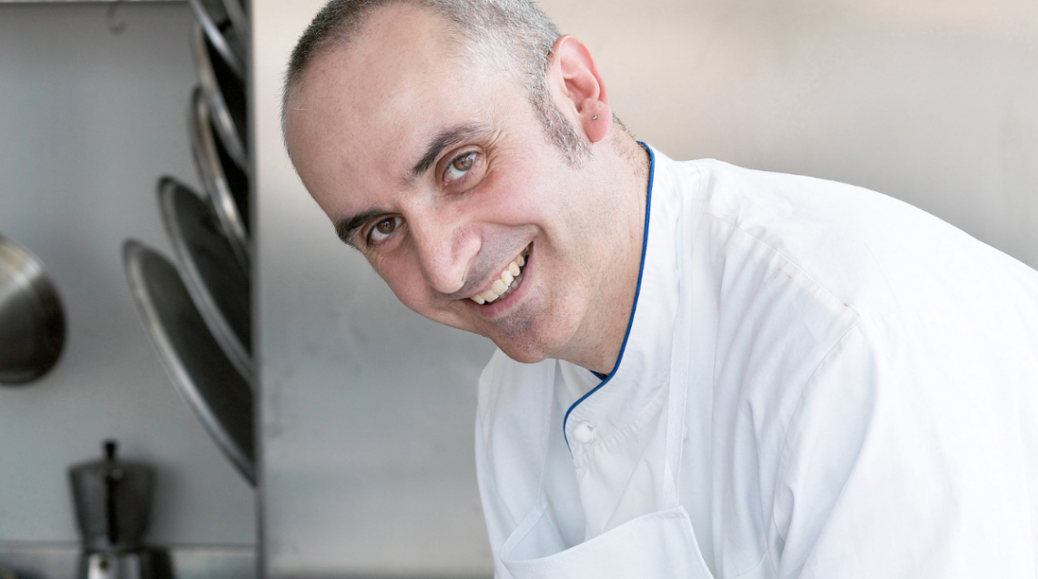 Ritratto dello chef Lazzaro Cimadoro sorridente, con indosso la giacca bianca professionale, in un ambiente di cucina moderna