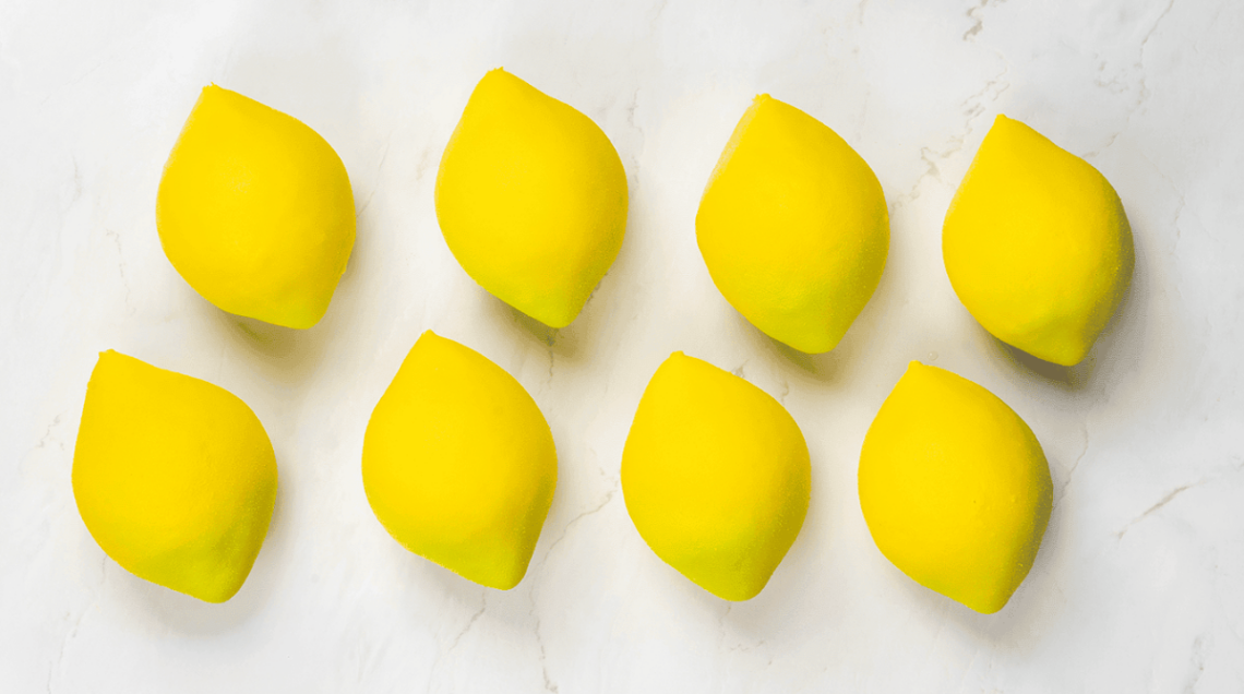 Serie di otto dolci a forma di limone giallo brillante disposti su una superficie di marmo.
