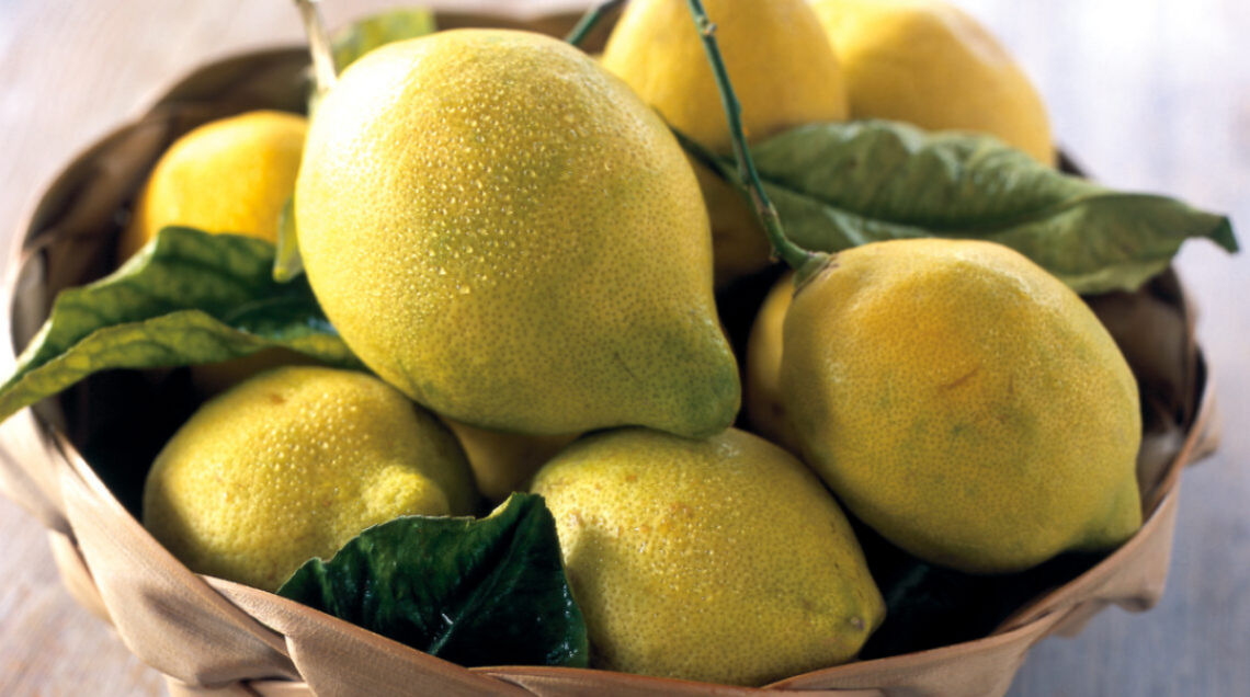 Un cesto di vimini colmo di limoni gialli e verdi appena raccolti, con foglie ancora attaccate ai rami.