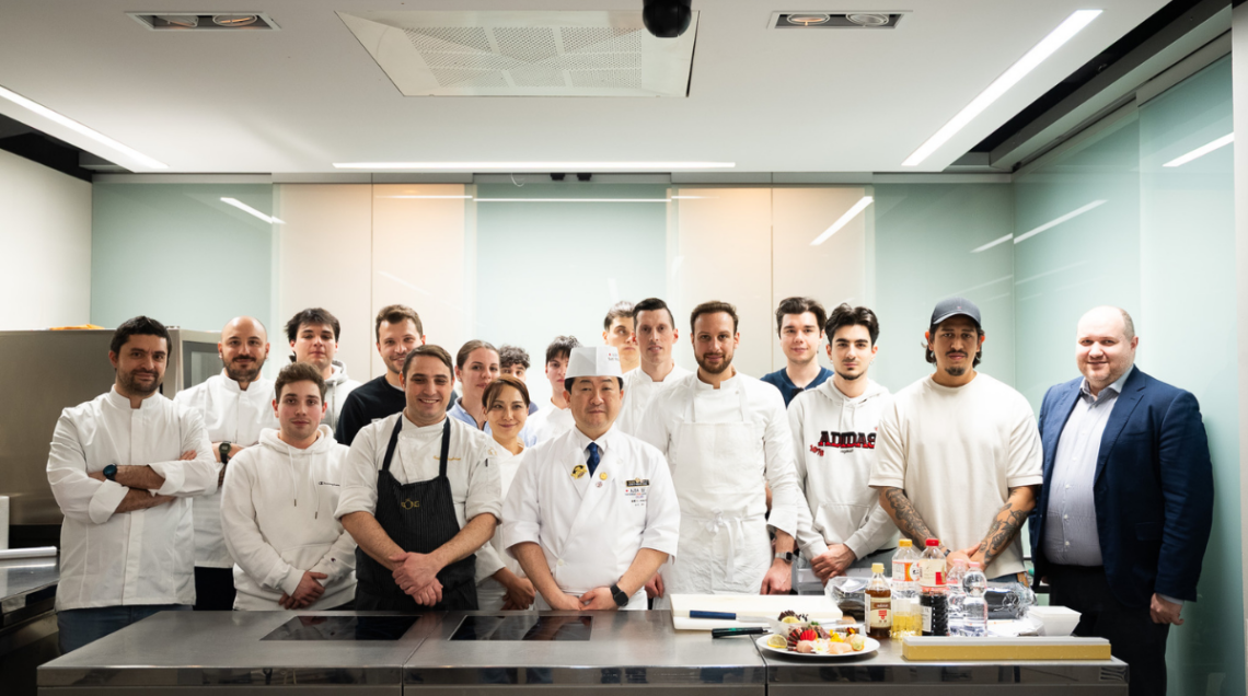 Foto di gruppo di una masterclass di cucina con lo chef istruttore al centro e i partecipanti schierati dietro un bancone professionale