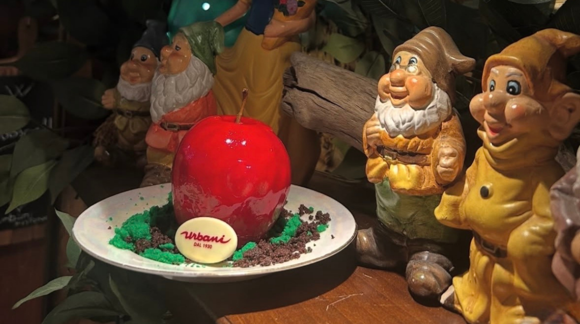 Dessert a forma di mela rossa lucida su piatto decorato, circondato da statuine dei sette nani.