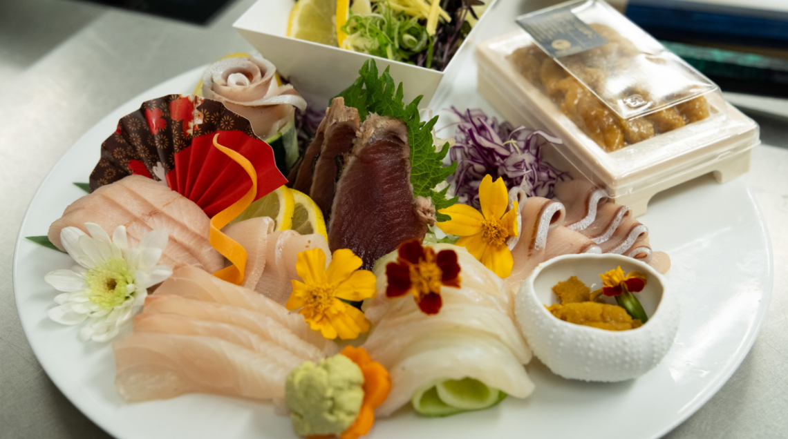 Piatto tondo bianco con una composizione artistica di sashimi, fiori eduli, wasabi e polpa di riccio di mare.