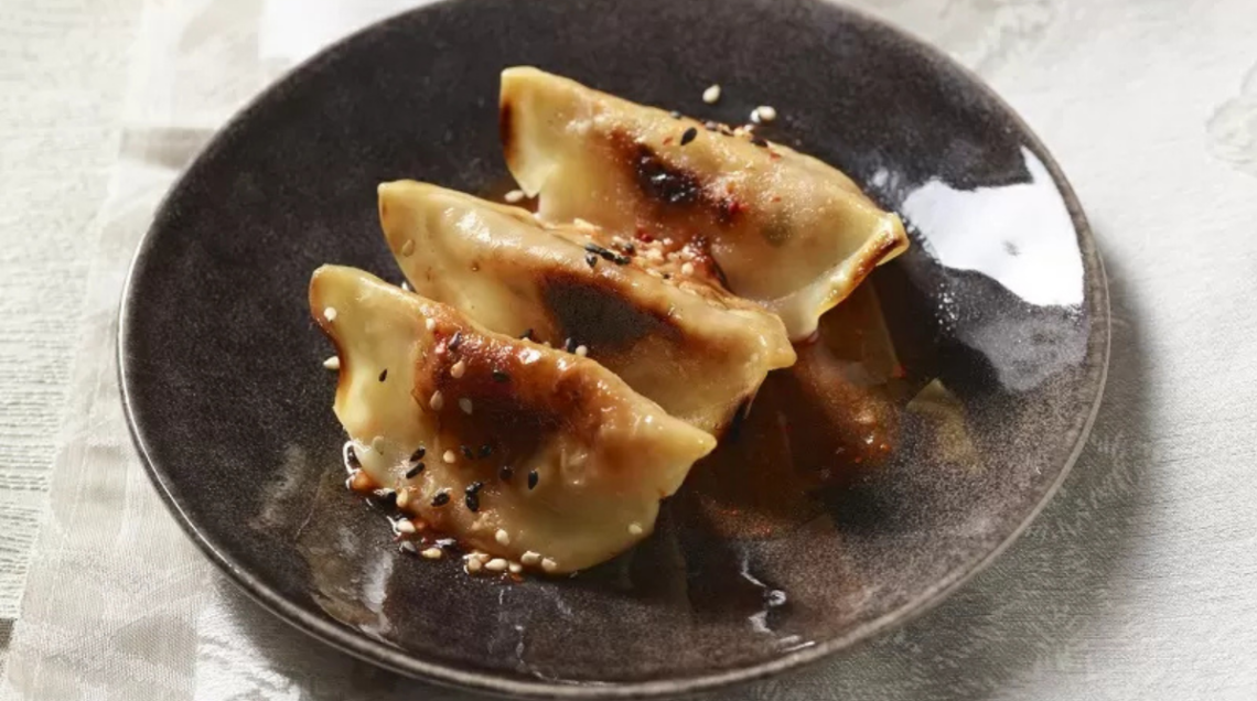 Tre ravioli alla piastra (mandu) serviti su un piatto scuro, guarniti con semi di sesamo nero e bianco e una salsa lucida.