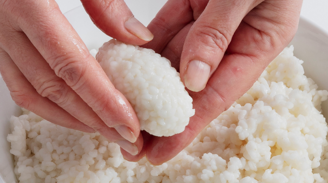 Mani che modellano con cura una piccola porzione di riso per sushi (nigiri) sopra una ciotola di riso bianco cotto