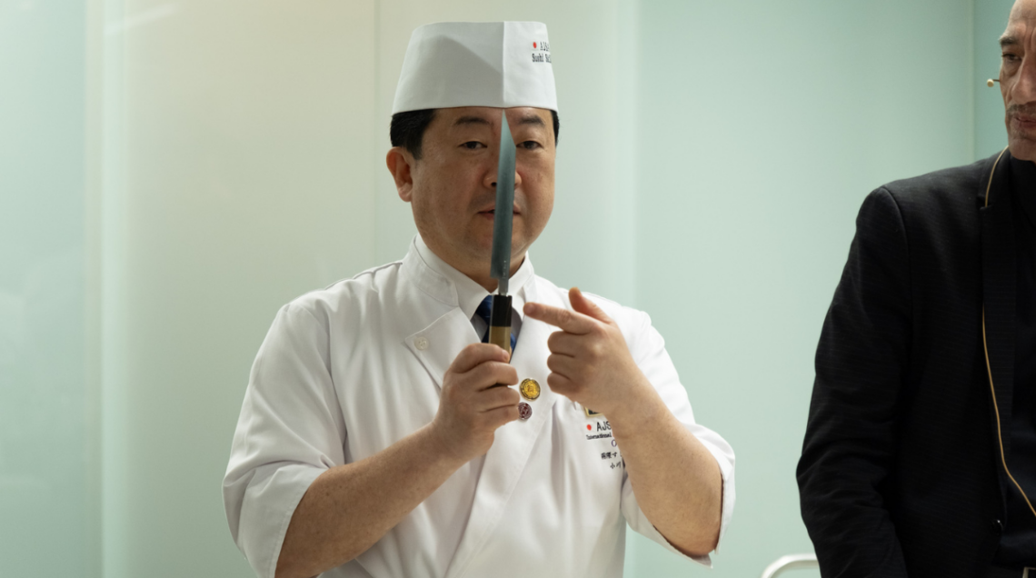 Il Maestro Hirotoshi Ogawache mostra verticalmente un coltello professionale da sushi, indicandone la lama con precisione