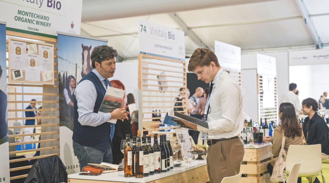 Visitatori e produttori presso lo stand Vinitaly Bio a Veronafiere durante l'edizione 2026, con esposizione di bottiglie di vino biologico toscano e brochure informative