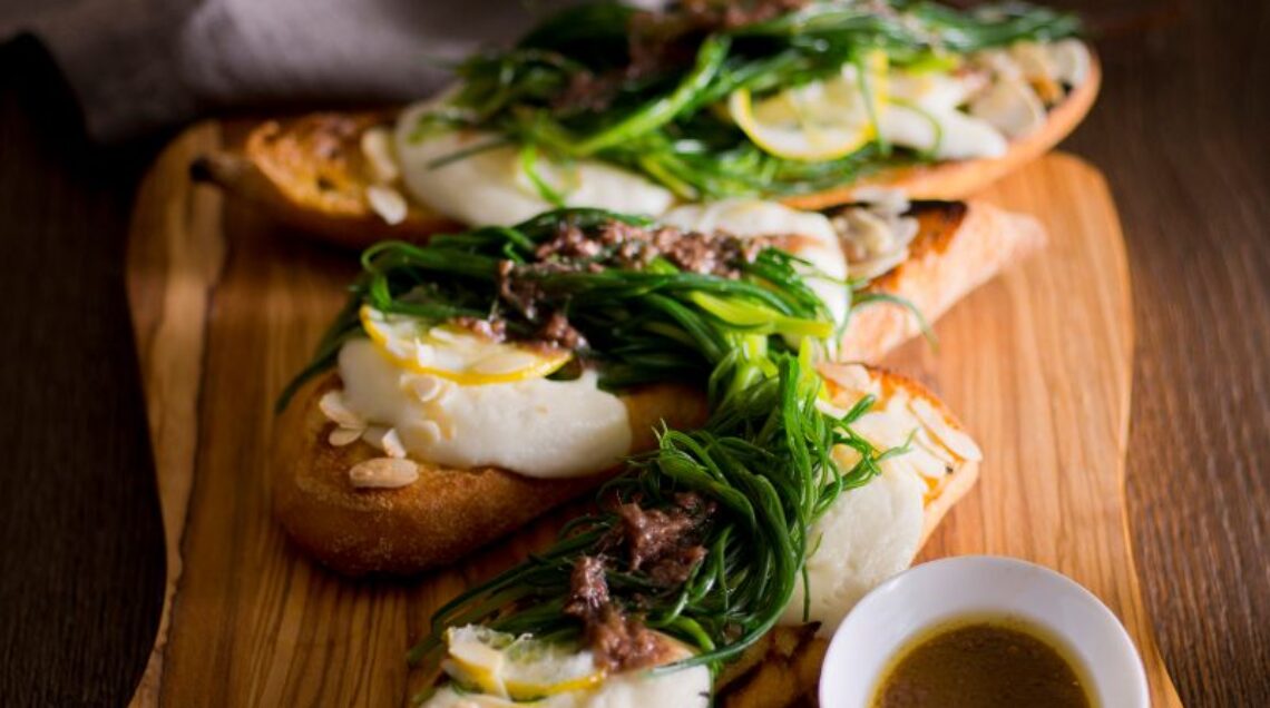 fette di baguette con agretti, bufala e alici, adagiate su vassoio di legno disposto su un tavolo