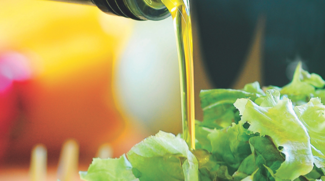Un filo di olio extravergine d'oliva dorato che scende da una bottiglia di vetro scuro su un'insalata verde fresca in una ciotola di vetro trasparente.
