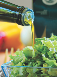 Un filo di olio extravergine d'oliva dorato che scende da una bottiglia di vetro scuro su un'insalata verde fresca in una ciotola di vetro trasparente.