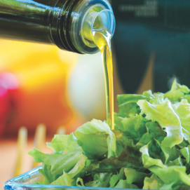 Un filo di olio extravergine d'oliva dorato che scende da una bottiglia di vetro scuro su un'insalata verde fresca in una ciotola di vetro trasparente.