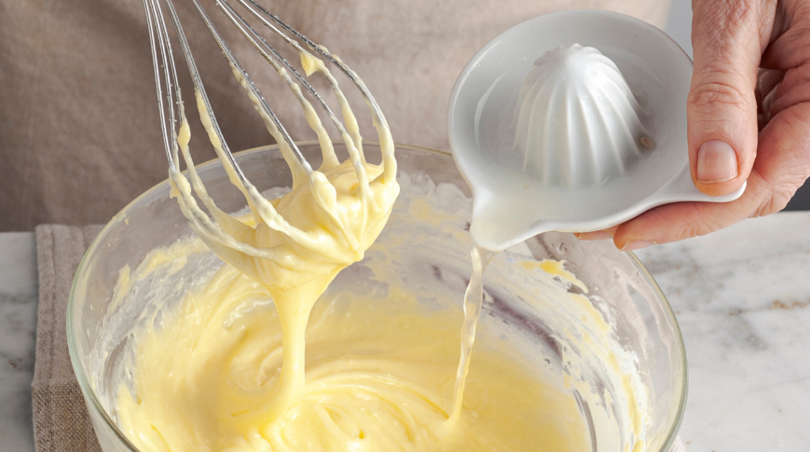 Una mano versa del succo di limone da uno spremiagrumi in ceramica bianca dentro una ciotola di vetro, mentre l'altra mano solleva una frusta da cucina ricoperta di maionese densa e cremosa.