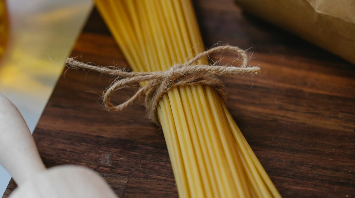 Un mazzo di spaghetti crudi legato con uno spago di iuta su un tagliere di legno scuro, accanto a un sacchetto di farina aperto e a un matterello in legno.