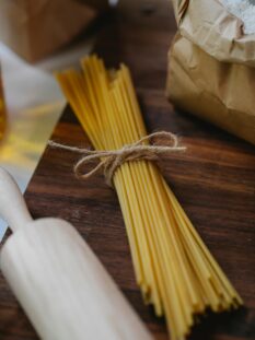Un mazzo di spaghetti crudi legato con uno spago di iuta su un tagliere di legno scuro, accanto a un sacchetto di farina aperto e a un matterello in legno.