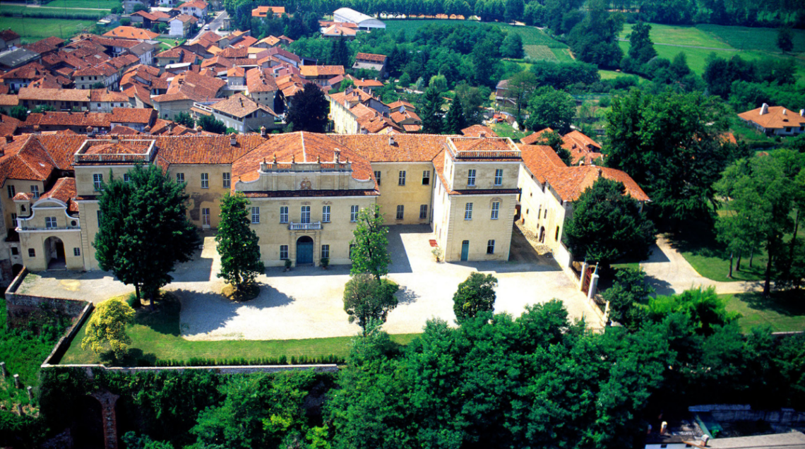 Veduta aerea del Castello di San Giorgio Canavese con il suo ampio cortile, i giardini circostanti e i tetti in coppi rossi del centro storico.