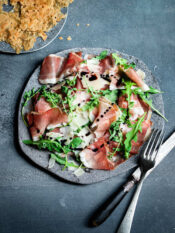 piatto di fette di speck, scaglie di grana, foglie di rucola e cialde di segale