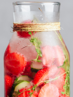 Bottiglia in vetro con acqua aromatizzata fredda, fragole a fette, cetrioli e rametti di timo.