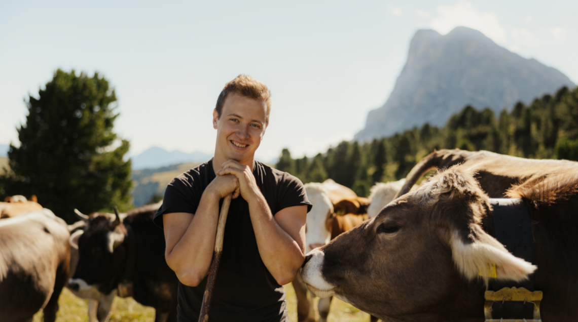 Giovane agricoltore sorridente tra le mucche al pascolo in un prato di montagna delle Dolomiti.