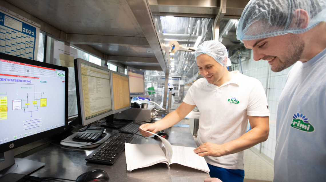 Due tecnici in camice bianco e cuffia monitorano i dati di produzione su schermi digitali in un ufficio tecnico.