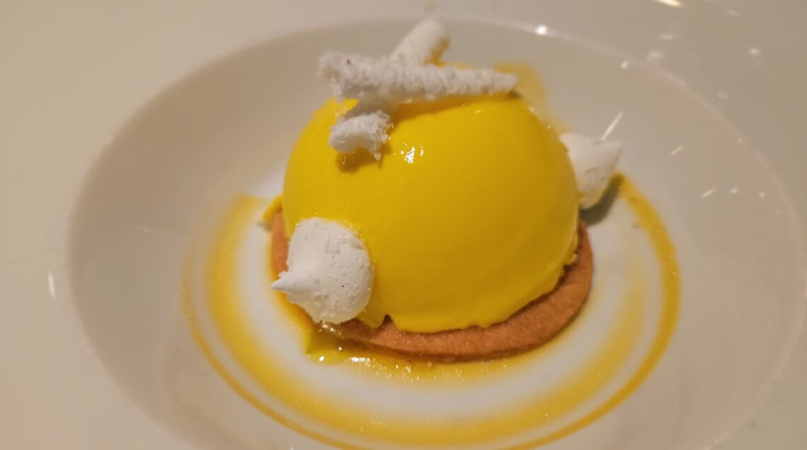 Dessert gourmet composto da una cupola di mousse gialla brillante su una base di dacquoise, decorata con piccole meringhe bianche e una riduzione di passion fruit circolare nel piatto.