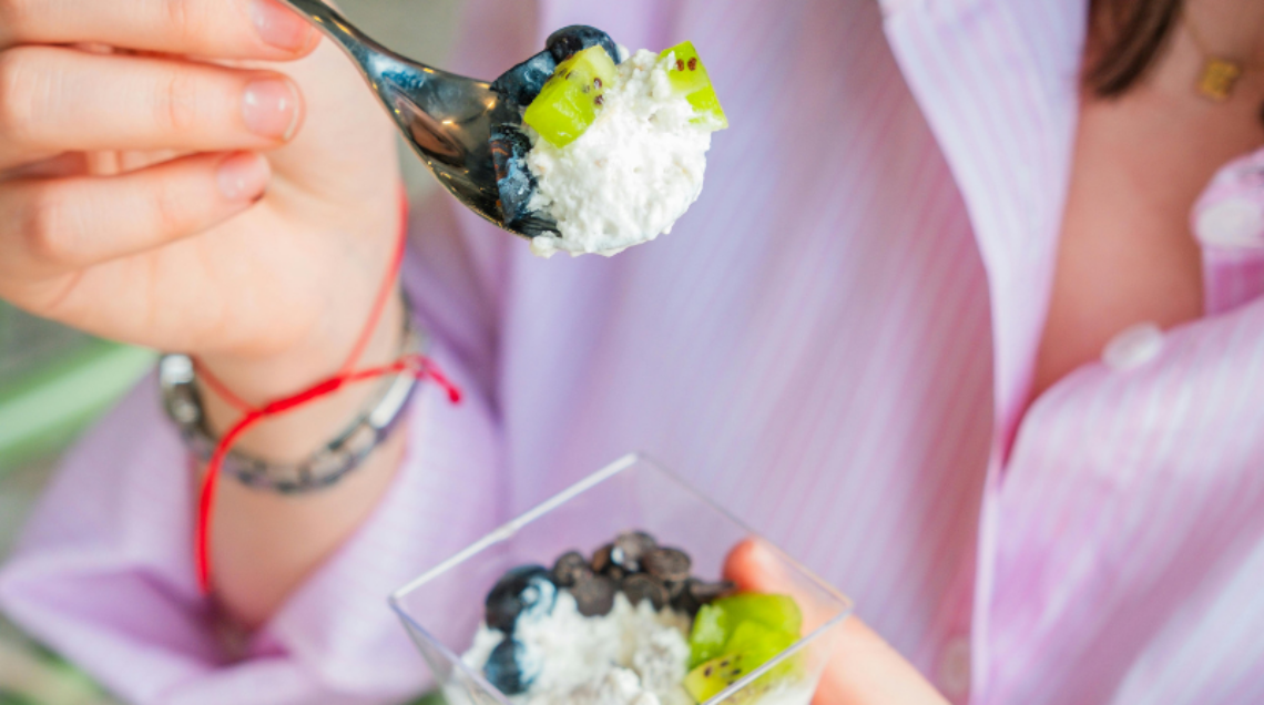 Una giovane donna con una camicia a righe rosa mentre mangia una ciotola di yogurt o ricotta con kiwi freschi, mirtilli e gocce di cioccolato, tenendo un cucchiaino sollevato.