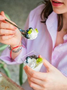 Una giovane donna con una camicia a righe rosa mentre mangia una ciotola di yogurt o ricotta con kiwi freschi, mirtilli e gocce di cioccolato, tenendo un cucchiaino sollevato.