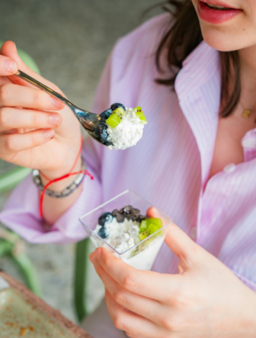 Una giovane donna con una camicia a righe rosa mentre mangia una ciotola di yogurt o ricotta con kiwi freschi, mirtilli e gocce di cioccolato, tenendo un cucchiaino sollevato.