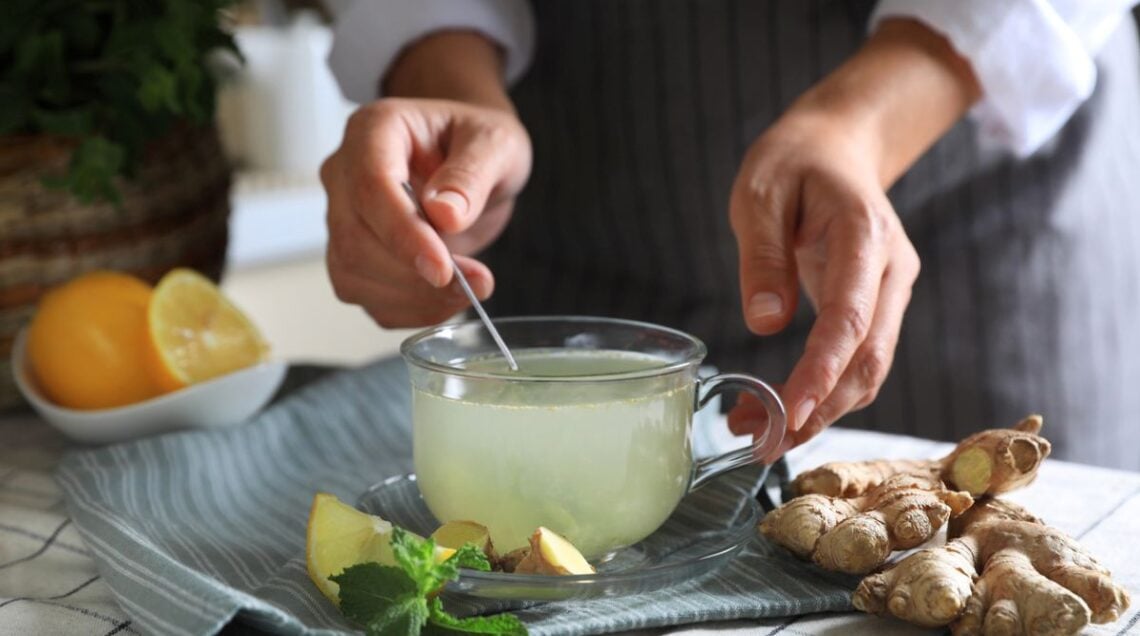 persona di cui si vedono solo le mani che mescola un infuso in una tazza di vetro trasparente appoggiata su un tavolo, con intorno limone tagliato a fette, zenzero in pezzi e foglioline di menta