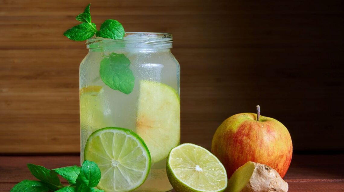vasetto di vetro contenente infuso di mela, limone, zenzero, guarnito con foglioline di menta, appoggiato su un tavolo con intorno una mela, un pezzo di zenzero, una fetta di lime e della menta