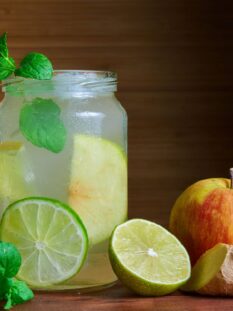 vasetto di vetro contenente infuso di mela, limone, zenzero, guarnito con foglioline di menta, appoggiato su un tavolo con intorno una mela, un pezzo di zenzero, una fetta di lime e della menta