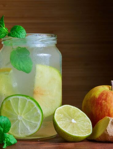 vasetto di vetro contenente infuso di mela, limone, zenzero, guarnito con foglioline di menta, appoggiato su un tavolo con intorno una mela, un pezzo di zenzero, una fetta di lime e della menta