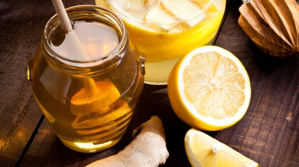 tavolo di legno visto dall'alto con sopra una caraffa contenende una bevanda dal colore giallo brillante, un vasetto di miele con dentro un coglimiele, circondati da limoni tagliati a fette e pezzi di zenzero