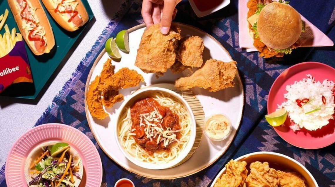 Tavola imbandita con piatti iconici di Jollibee: pollo fritto Chickenjoy, spaghetti dolci con salsiccia, hamburger e riso bianco. 