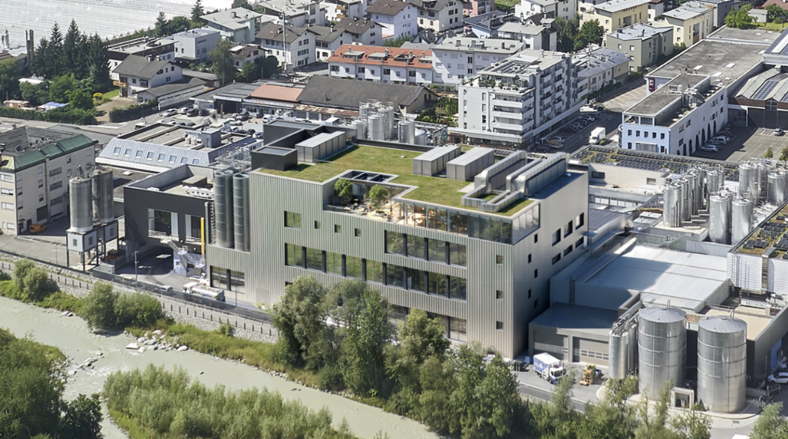 Veduta aerea del moderno stabilimento Brimi a Bressanone, con tetto verde e silos, situato vicino a un fiume.