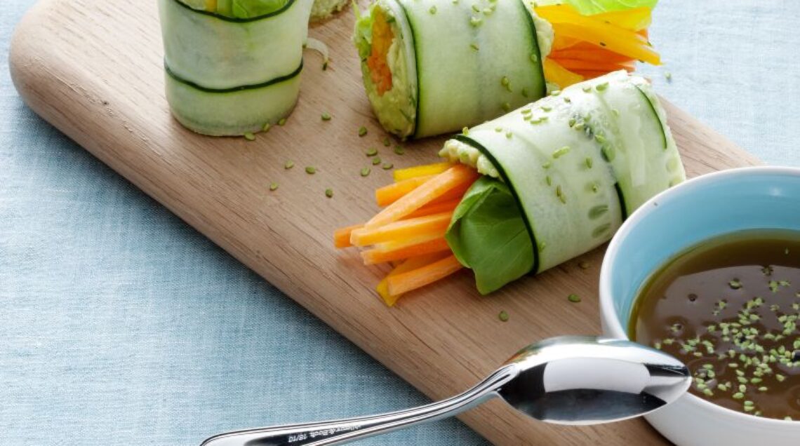 rotolini di cetriolo con crema di avocado e verdure disposti su un vassoio di legno, con salsina a parte