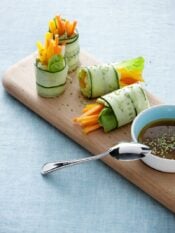 rotolini di cetriolo con crema di avocado e verdure disposti su un vassoio di legno, con salsina a parte