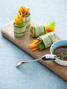 rotolini di cetriolo con crema di avocado e verdure disposti su un vassoio di legno, con salsina a parte