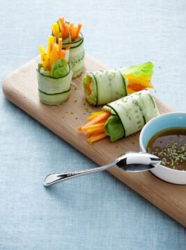 rotolini di cetriolo con crema di avocado e verdure disposti su un vassoio di legno, con salsina a parte