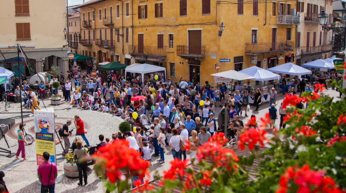 Una folla di persone riunita nella piazza principale di San Giorgio Canavese tra stand bianchi, palloncini e fiori rossi in primo piano.